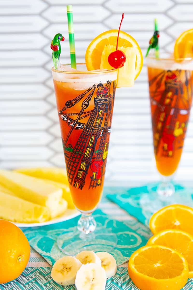 Rum Runner Cocktail in a tall pirate designed pilsner glass on a turquoise tablecloth surrounded by oranges, pineapple wedges and banana slices.
