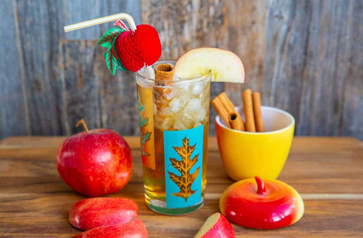 Apple Cider Cocktail in a turquoise and gold glass on a wood board surrounded by cinnamon sticks and apples.