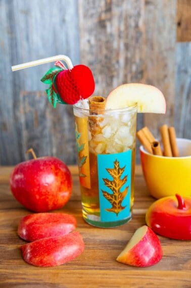 Apple Cider Cocktail in a turquoise and gold glass on a wood board surrounded by cinnamon sticks and apples.