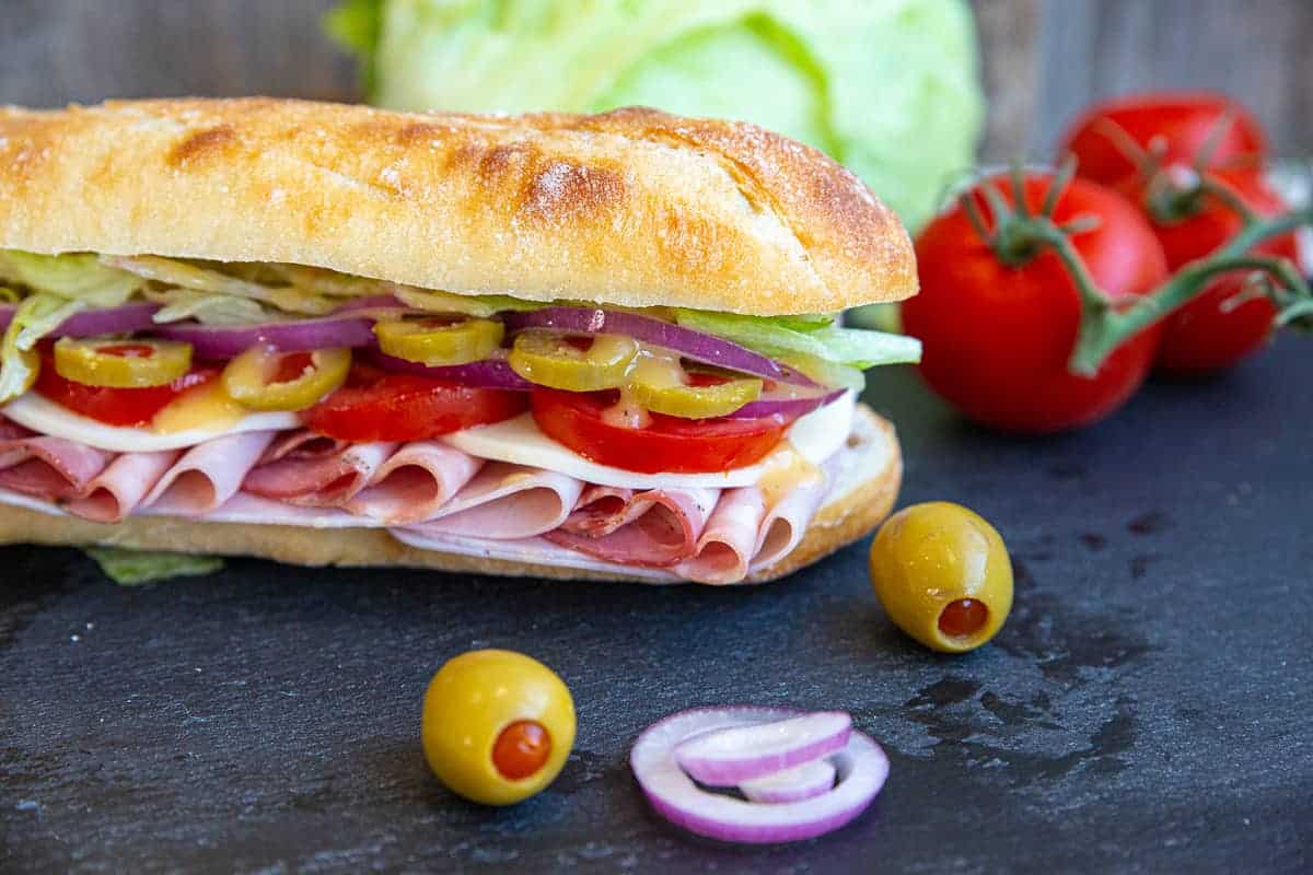 Close up of Italian Sub on a black slate board with olives, onion, tomatoes and lettuce surrounding it.