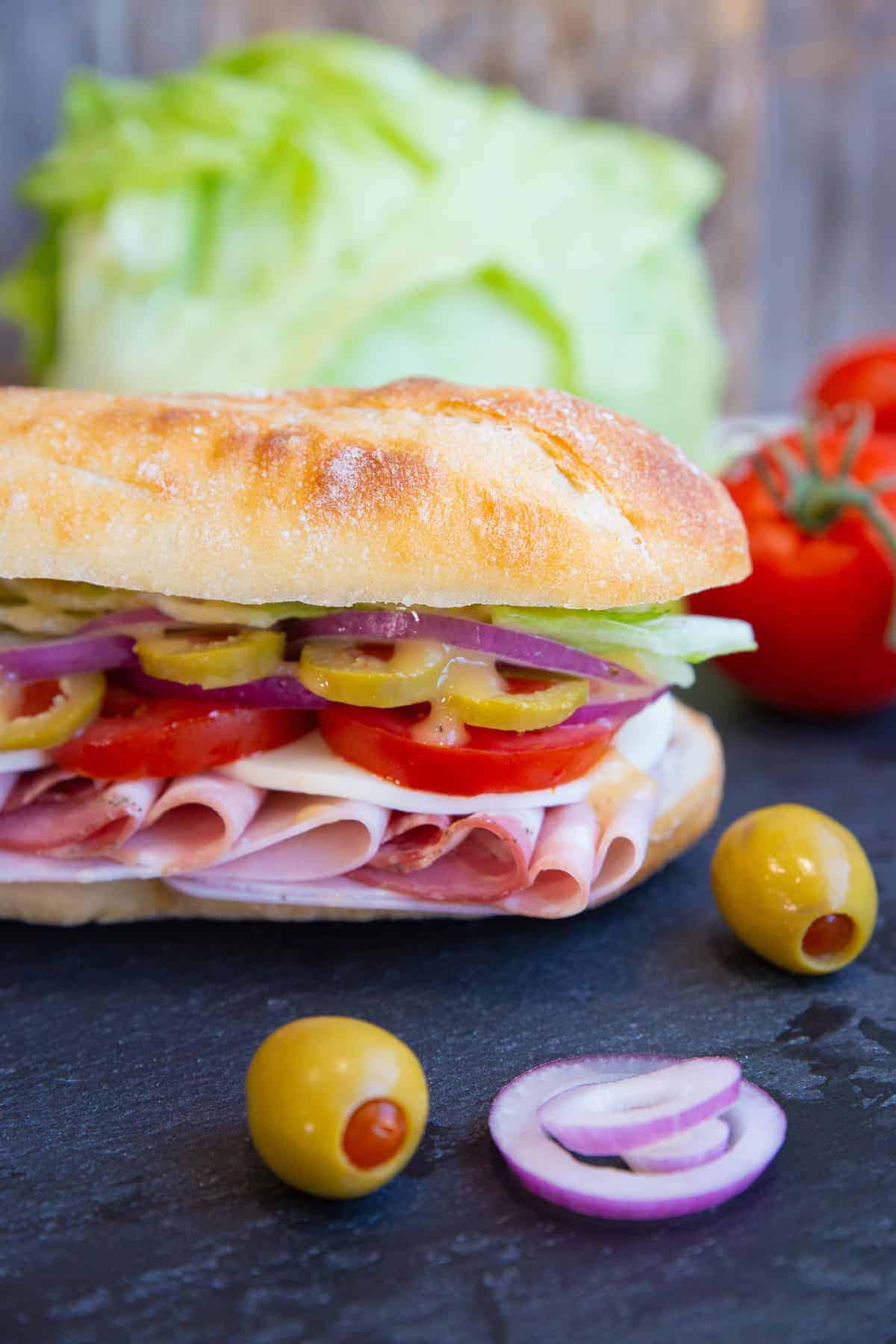 Close up of Italian Sub on a black slate board with olives, onion, tomatoes and lettuce surrounding it.