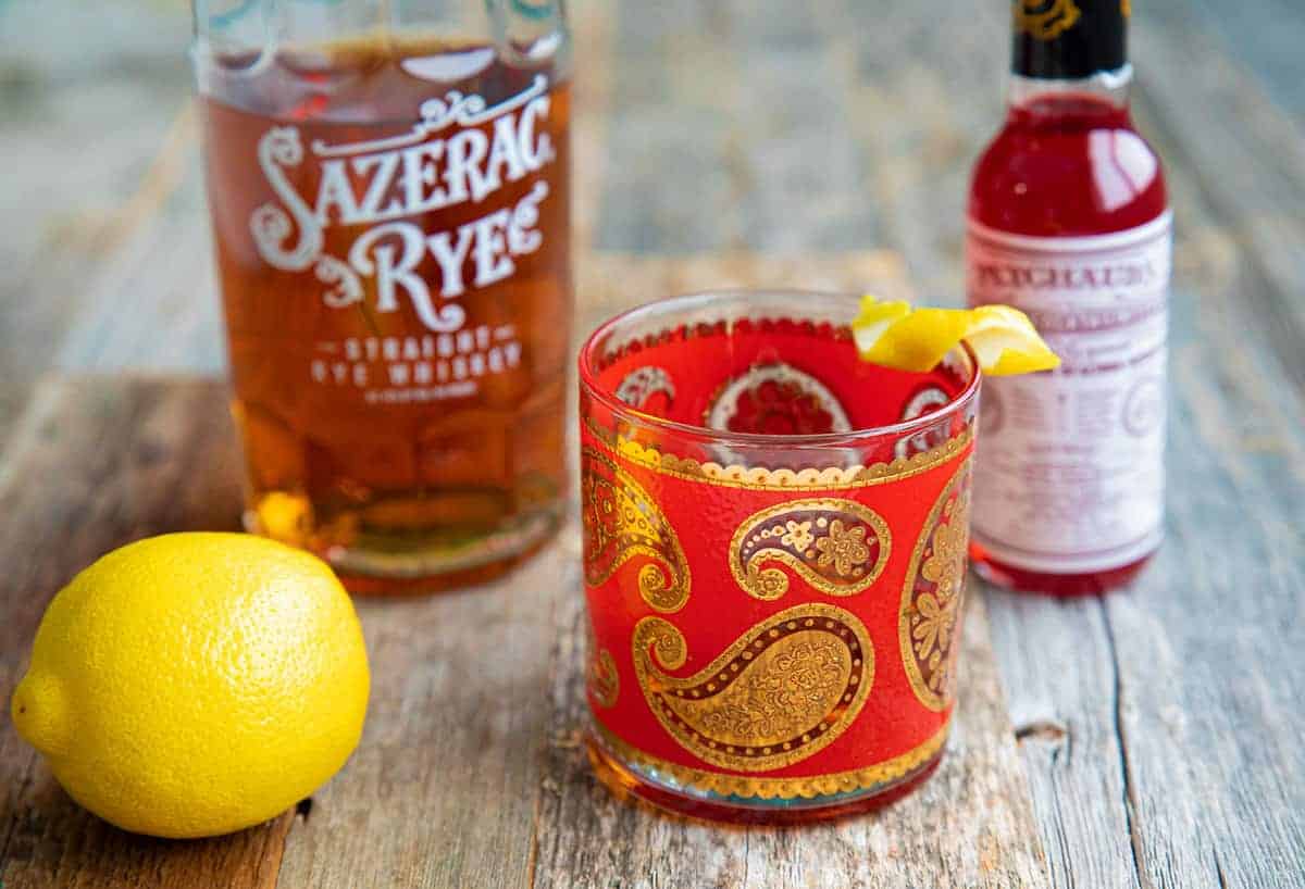 Sazerac Cocktail in a vintage old fashioned glass on wood with a lemon, bottle of whiskey and bottle of bitters.