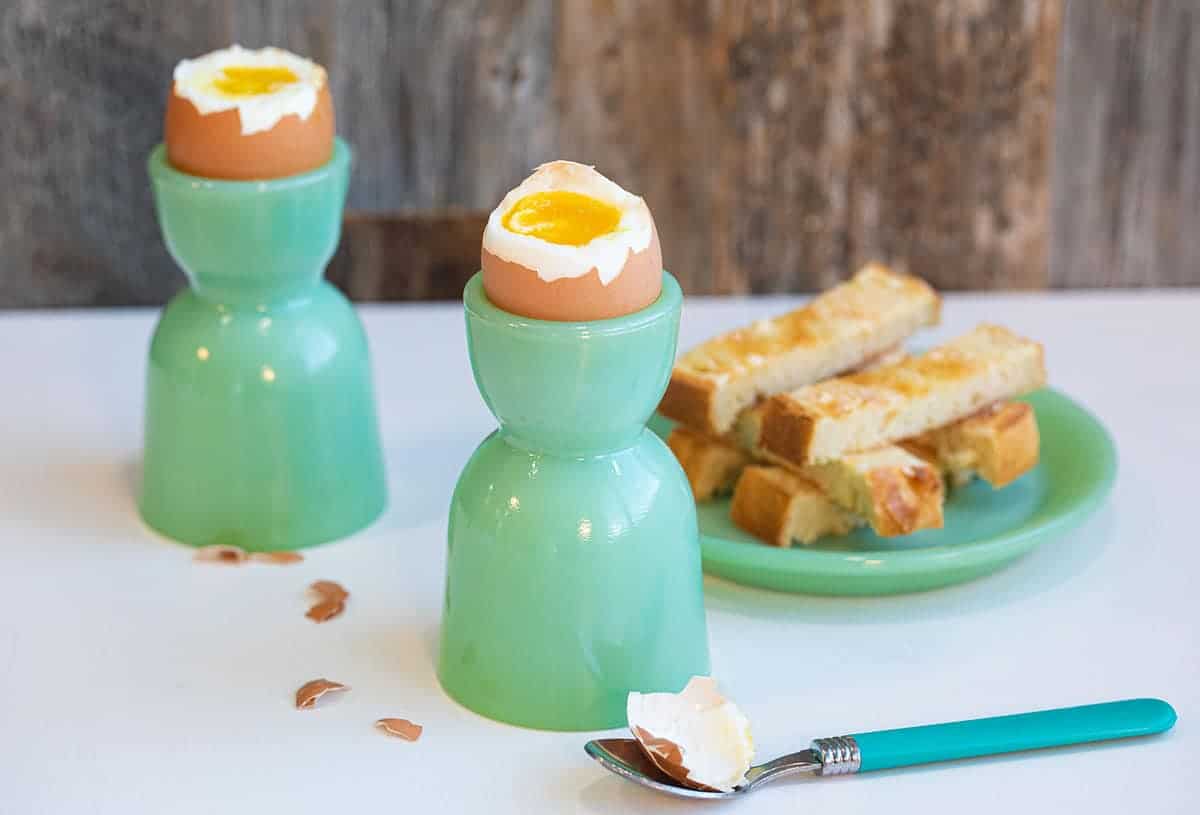 Soft boiled eggs with their tops removed, showing the inner yolk, next to some toast soldiers on jadeite kitchenware.