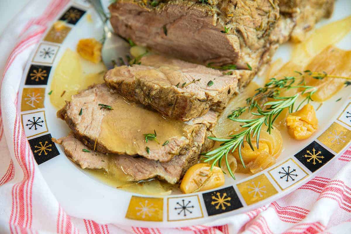 Close up of a sliced pork roast, covered in juices and sprinkled with rosemary, on a plate.