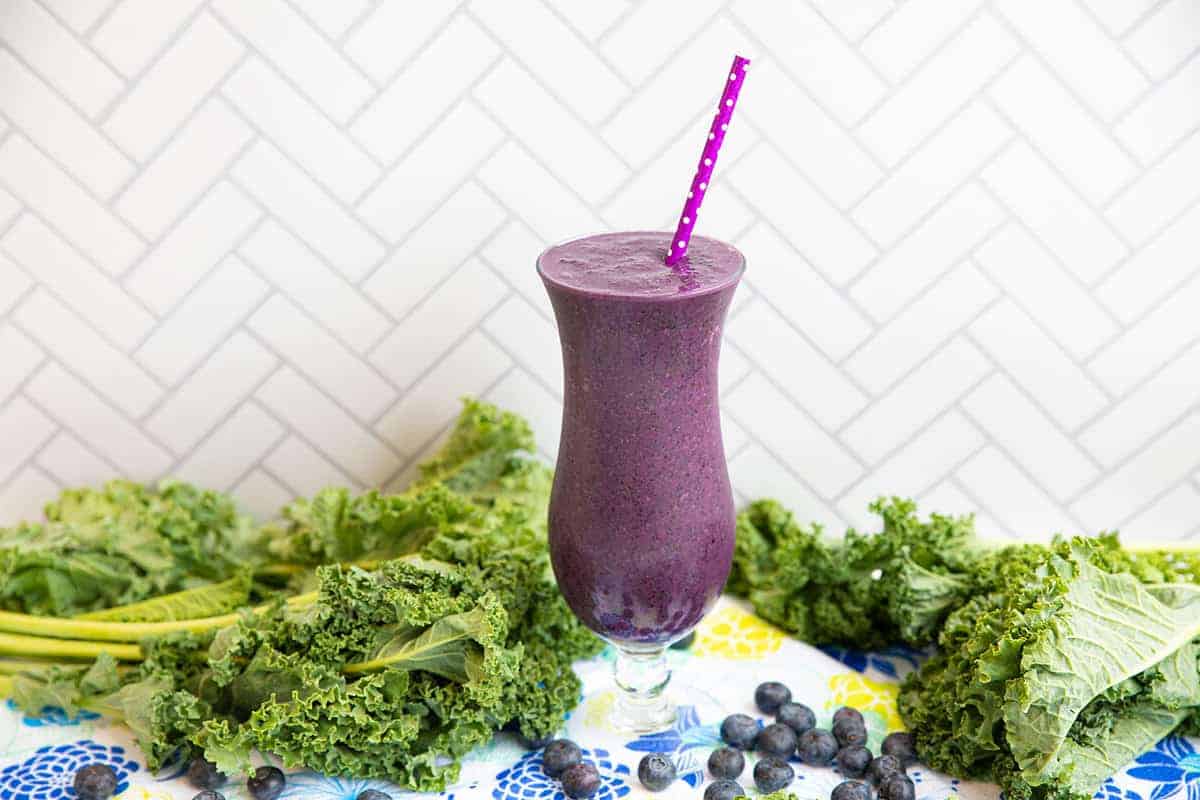 Blueberry Kale Smoothie on a white background surrounded by loose blueberries and bunches of kale.