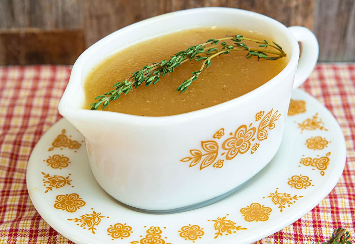 A serving bowl filled with pork gravy on a white plate, topped with a sprig of rosemary. 