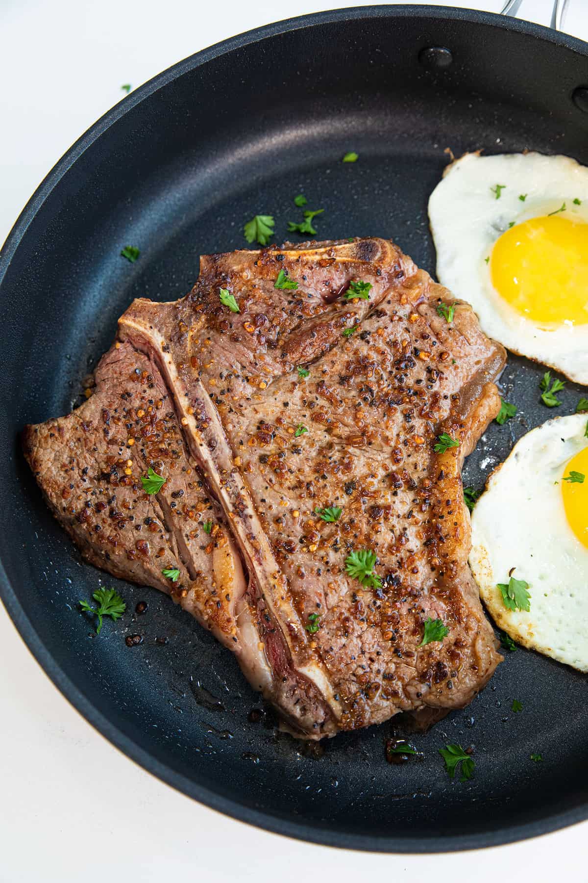 Close up of a T bone steak with two friend eggs next to it on a cast iron pan