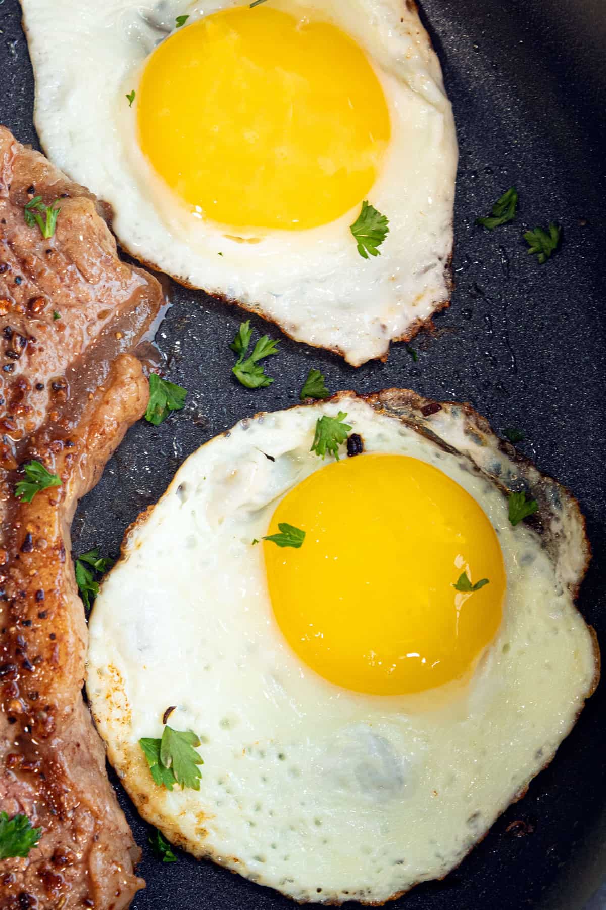 A close-up of two sunny side up fried eggs, next to the edge of a steak on a black plate, dotted with parsley