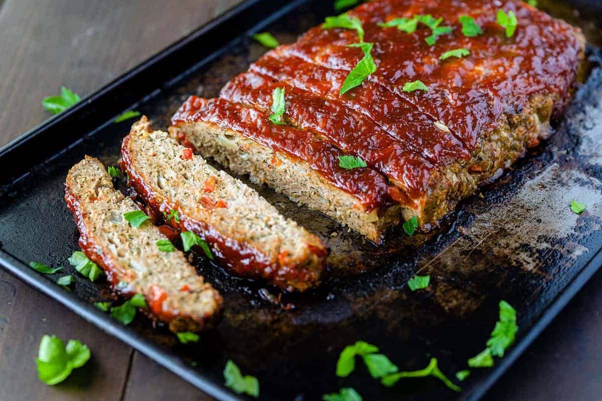 A sliced turkey meatloaf topped with cup up parsley on a black plate. 