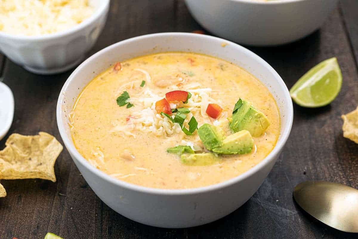 A bowl of finished white chicken chili, topped with avocado and peppers, on a wooden table