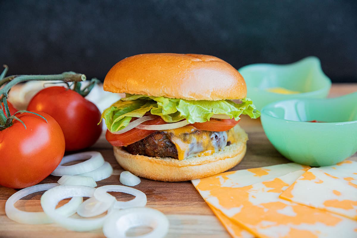 Air Fryer Hamburgers on a wood cutting board with tomatoes, onion, cheese and lettuce.