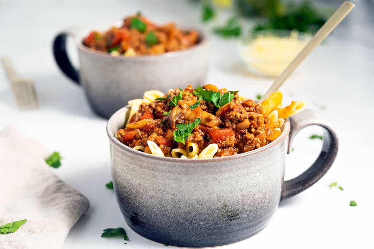 A cupful of penne pasta and meat sauce on a white backdrop. 