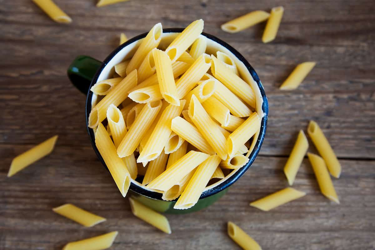A cup full of dried penne pasta on a wooden table.