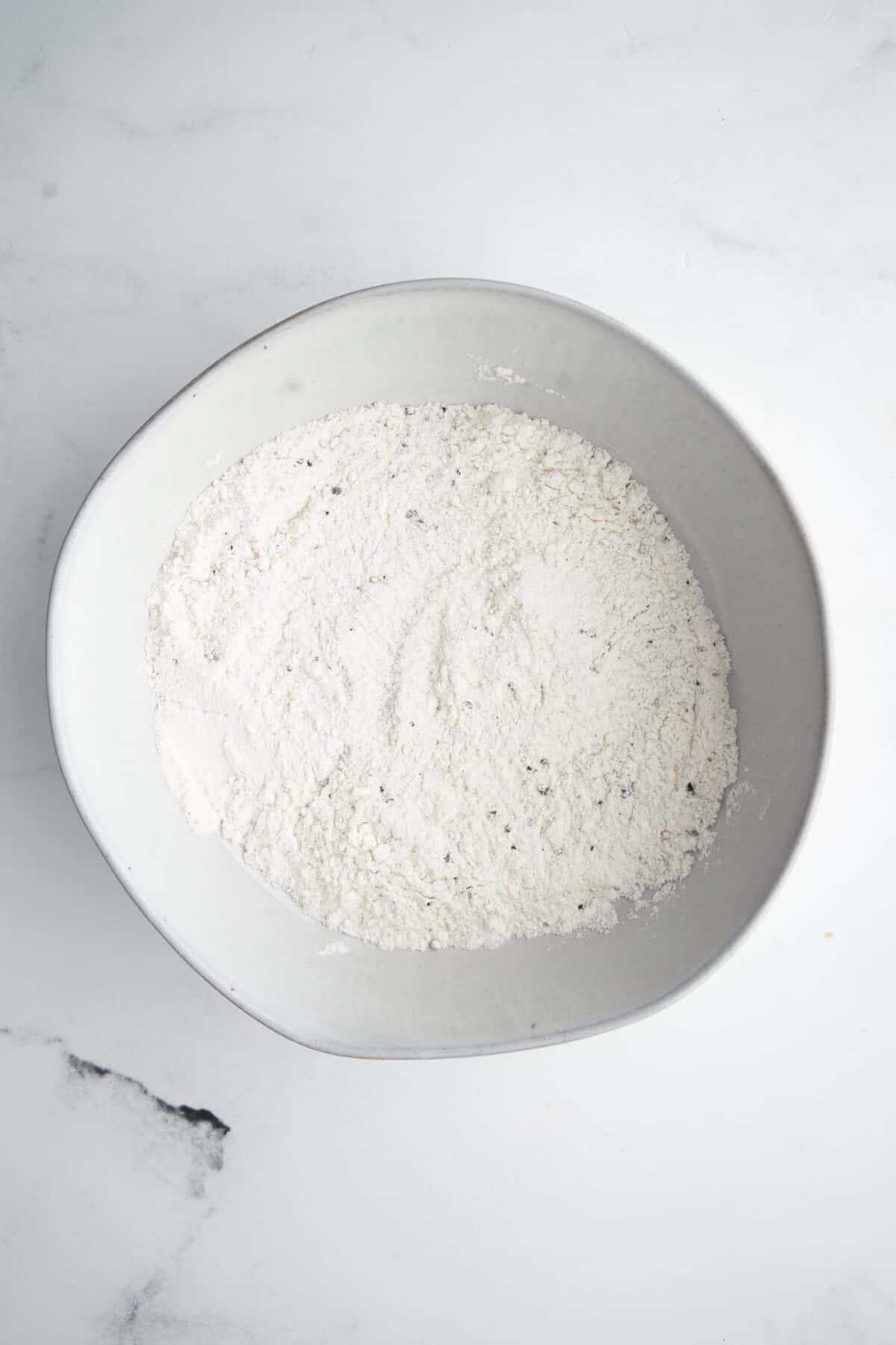 A white ceramic bowl filled with a mound of white flour sits on a white marble countertop.