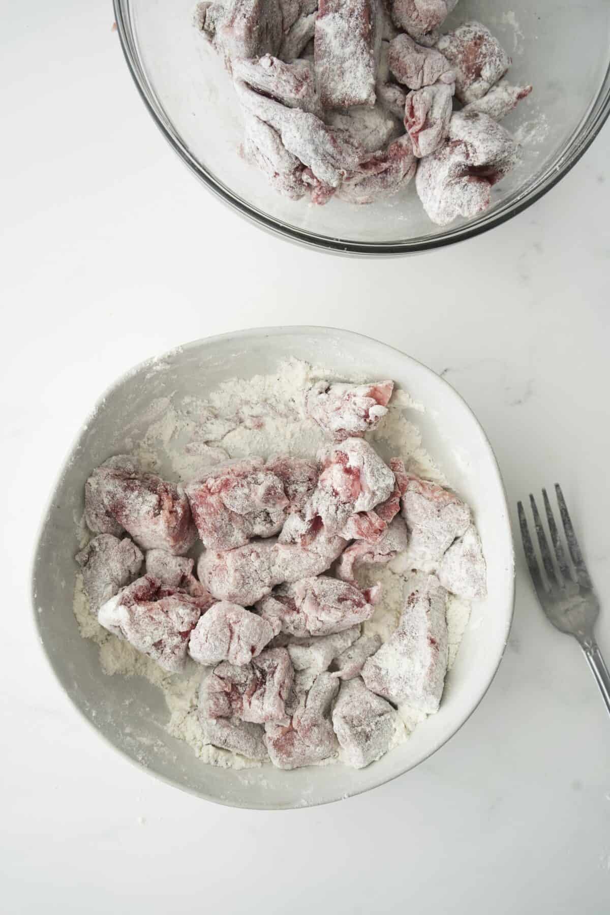 Raw beef pieces coated in flour in a white bowl, with a fork beside the bowl and a second bowl with more floured beef pieces partially visible at the top, all on a white surface.