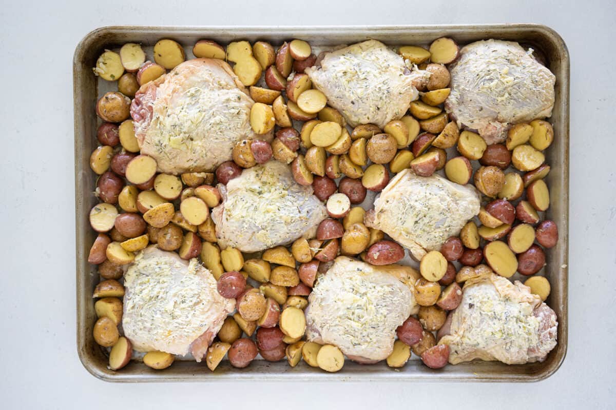 raw chicken thighs buttered up on and a baking sheet with potatoes 
