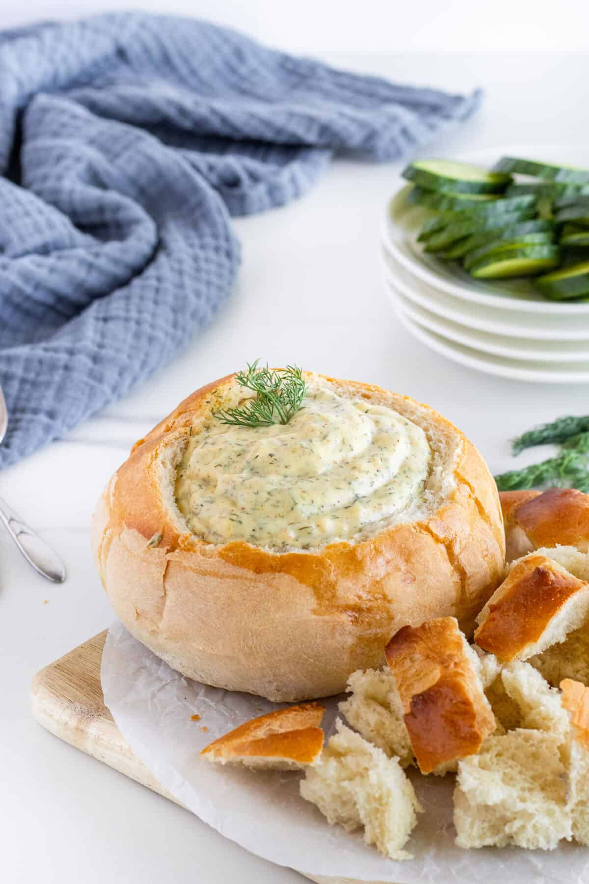 dill dip in a bread bowl with bread cubes and cucumbers 