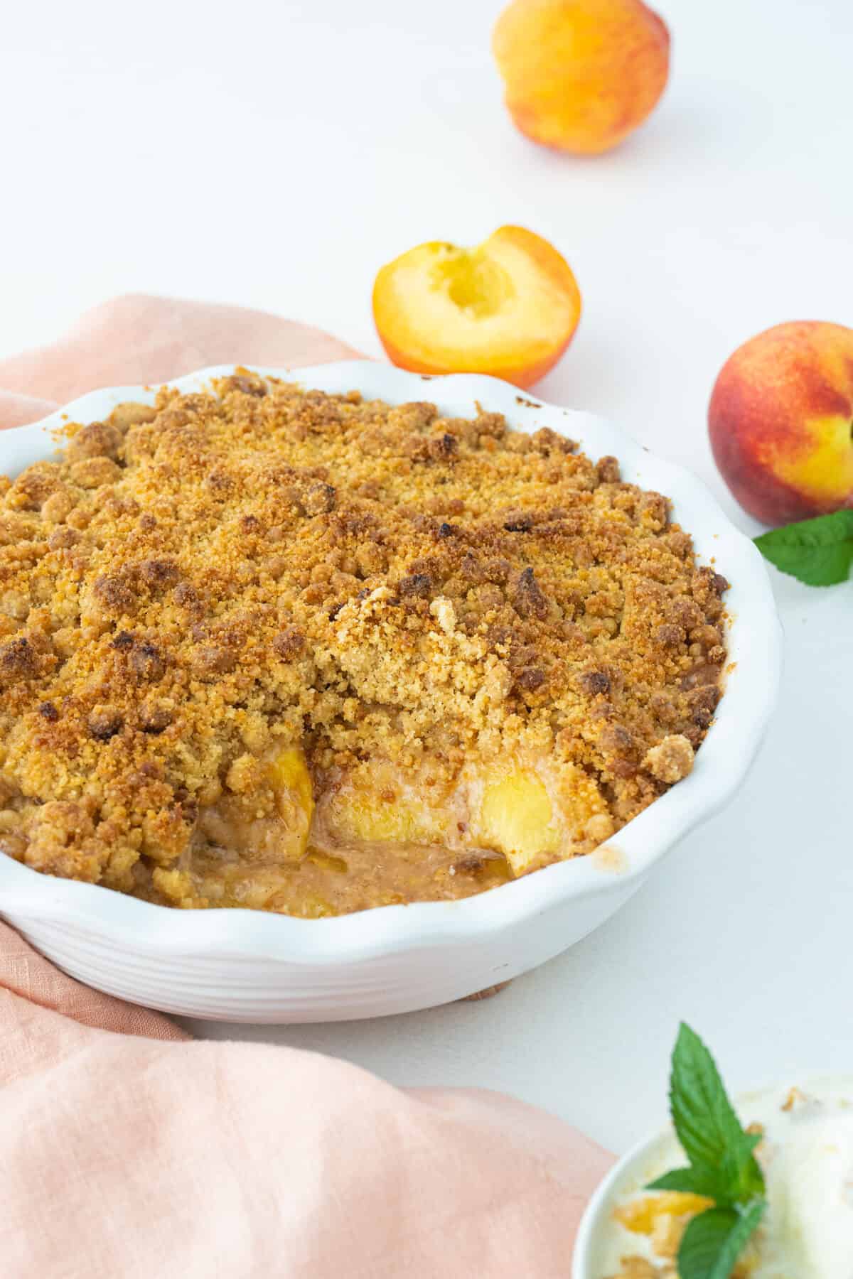 peach crumble in a white pie plate with fresh peaches on the counter beside it