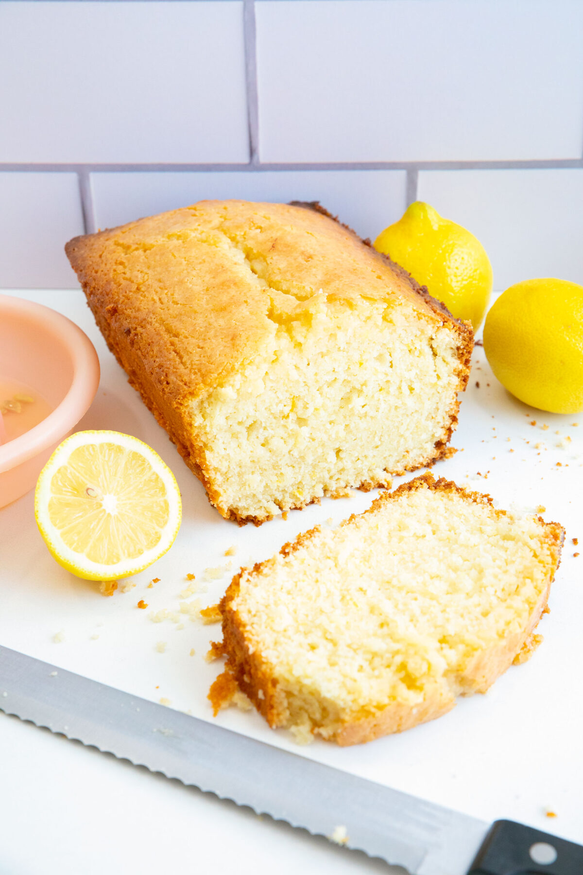 A loaf of lemon pound cake with one slice cut, surrounded by fresh lemons and a knife on a white surface, against a tiled background.