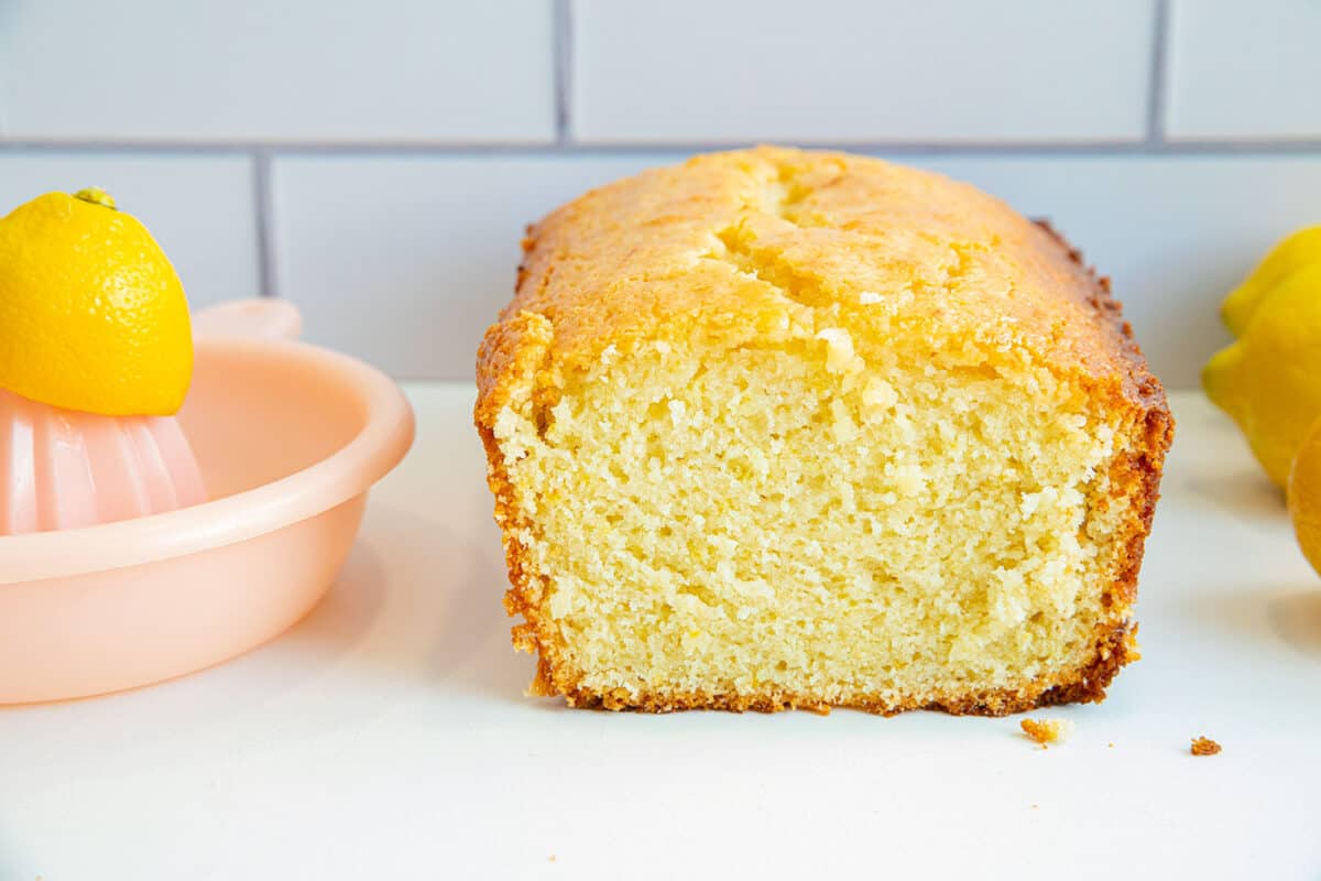 A sliced lemon pound cake loaf sits on a white surface, with a pink lemon squeezer and whole lemons beside it. The background features white subway tiles.