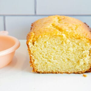 A sliced lemon pound cake loaf sits on a white surface, with a pink lemon squeezer and whole lemons beside it. The background features white subway tiles.