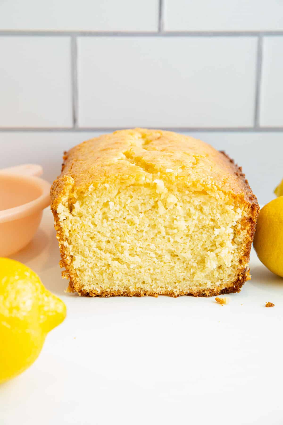 Sliced loaf of lemon pound cake on a white surface with fresh lemons and a pink bowl in the background, in front of a white tiled wall.