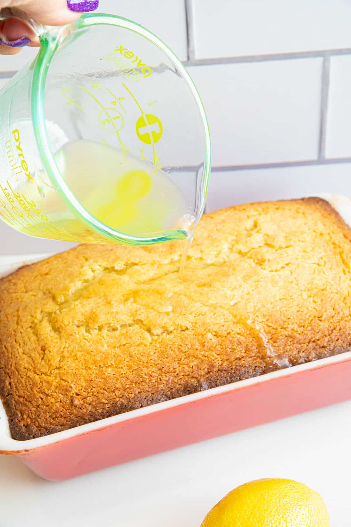 A hand pours lemon glaze from a glass measuring cup over a freshly baked lemon pound cake in a pink dish. A lemon sits nearby on the white counter.