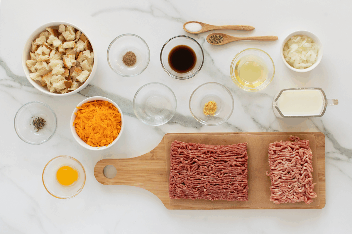 A variety of cheese stuffed meatloaf ingredients arranged on a white marble surface, including ground beef and pork, shredded cheese, bread cubes, egg, milk, chopped onions, seasonings, and sauces in bowls and spoons.