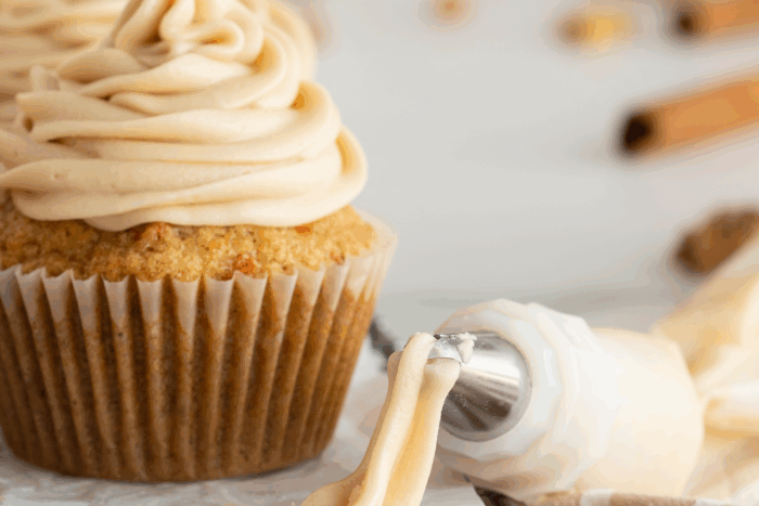 A close-up of a cupcake with light brown maple frosting, next to a piping bag with frosting on the tip. The background is blurred with hints of cinnamon sticks.