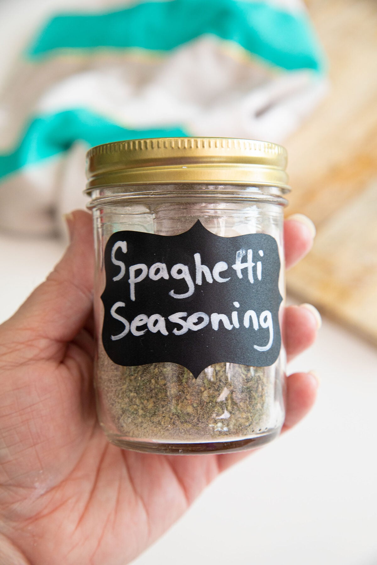 A hand holds a glass jar with a gold lid labeled Spaghetti Seasoning. The jar contains a greenish spice blend. A towel and cutting board are blurred in the background.
