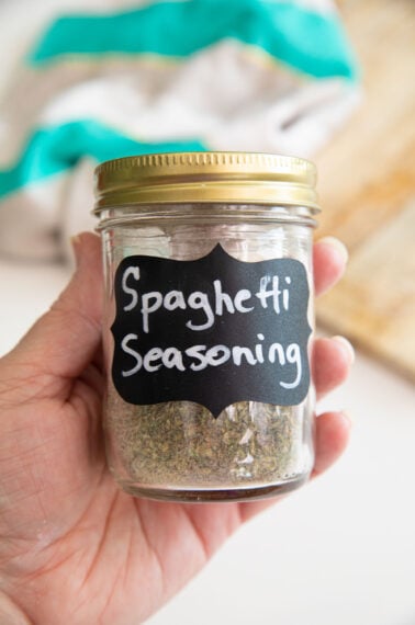 A hand holds a glass jar with a gold lid labeled Spaghetti Seasoning. The jar contains a greenish spice blend. A towel and cutting board are blurred in the background.