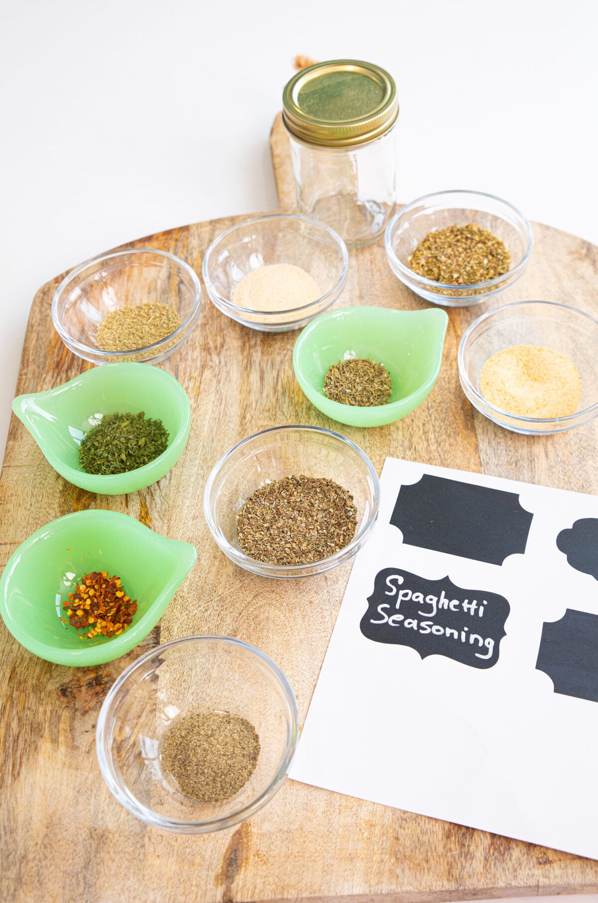 A wooden board with small bowls of various spices, a glass jar with a gold lid, and a sheet labeled Spaghetti Seasoning with blank sticker labels.