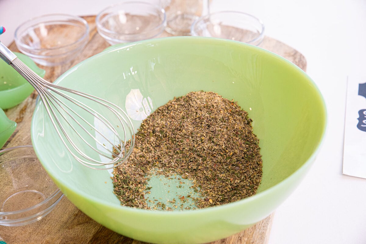 A green bowl containing a mix of dried herbs for spaghetti seasoning with a metal whisk resting inside. Small glass bowls and a wooden surface are visible in the background.