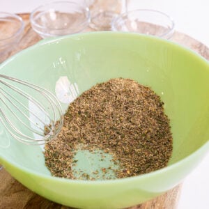 A green bowl containing a mix of dried herbs for spaghetti seasoning with a metal whisk resting inside. Small glass bowls and a wooden surface are visible in the background.