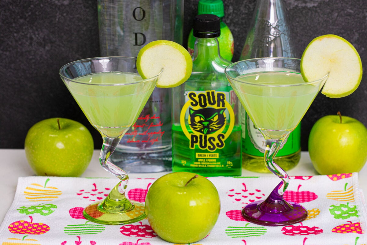 two appletini cocktails on a placement surrounded by green apples and the ingredients used to make them.