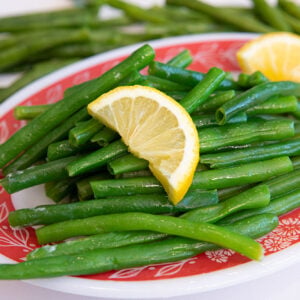 A plate of boiled green beans garnished with two lemon wedges, served on a red and white patterned dish, with more green beans in the background.
