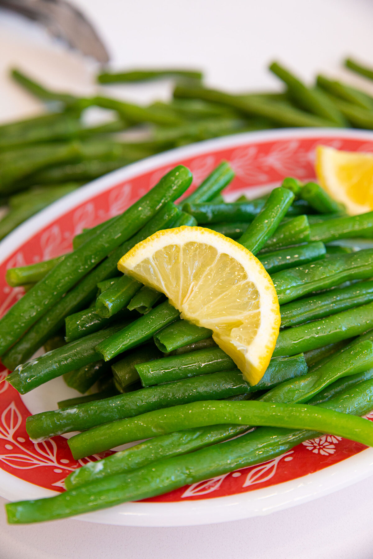 A plate of steamed green beans topped with a lemon wedge sits on a red and white patterned dish, with more green beans and a utensil blurred in the background.