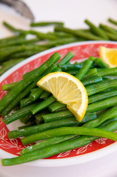 A plate of steamed green beans topped with a lemon wedge sits on a red and white patterned dish, with more green beans and a utensil blurred in the background.