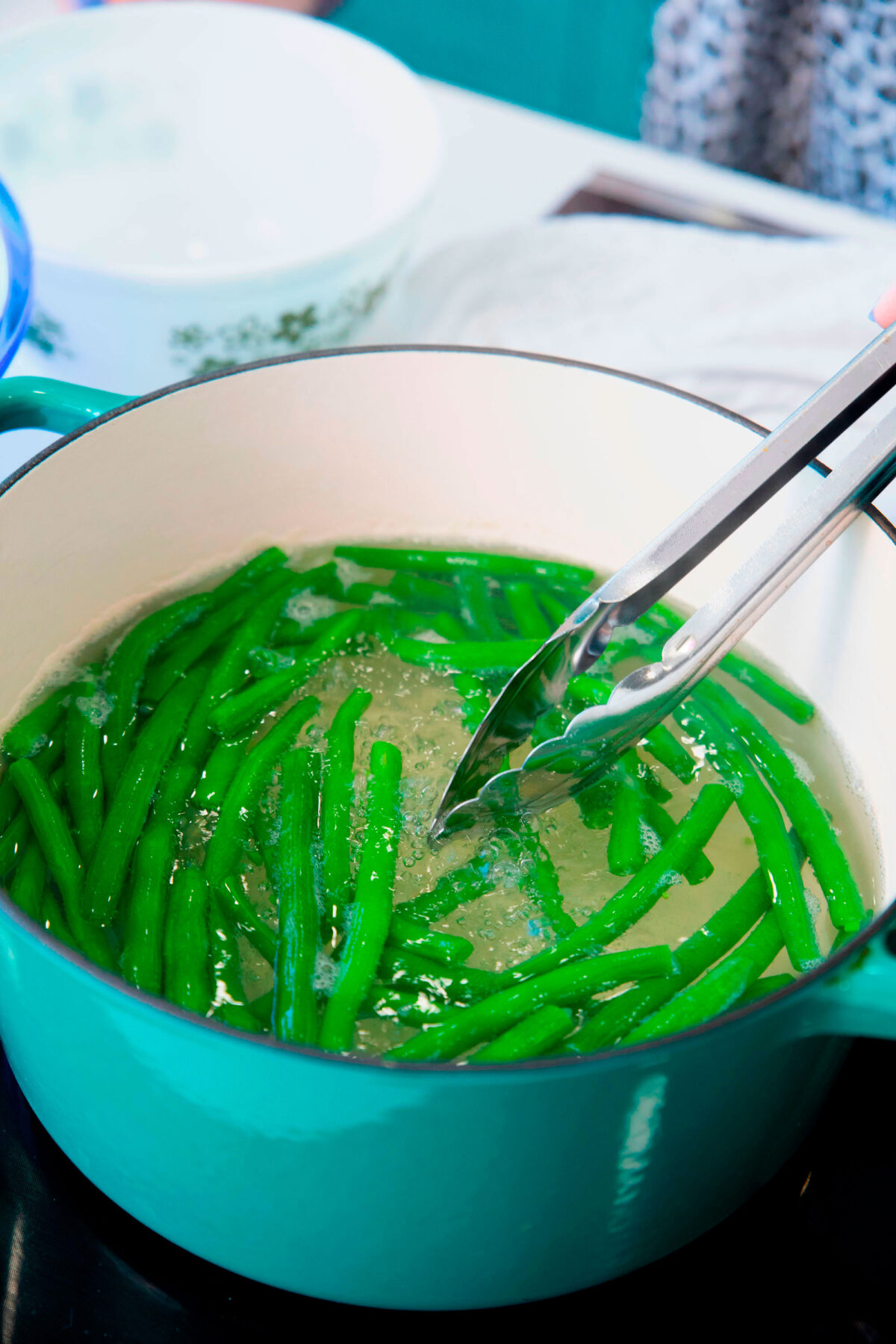 Boiled green beans are being blanched in a large turquoise pot, with metal tongs lifting some of the vibrant beans. A white bowl and towel can be seen in the background.