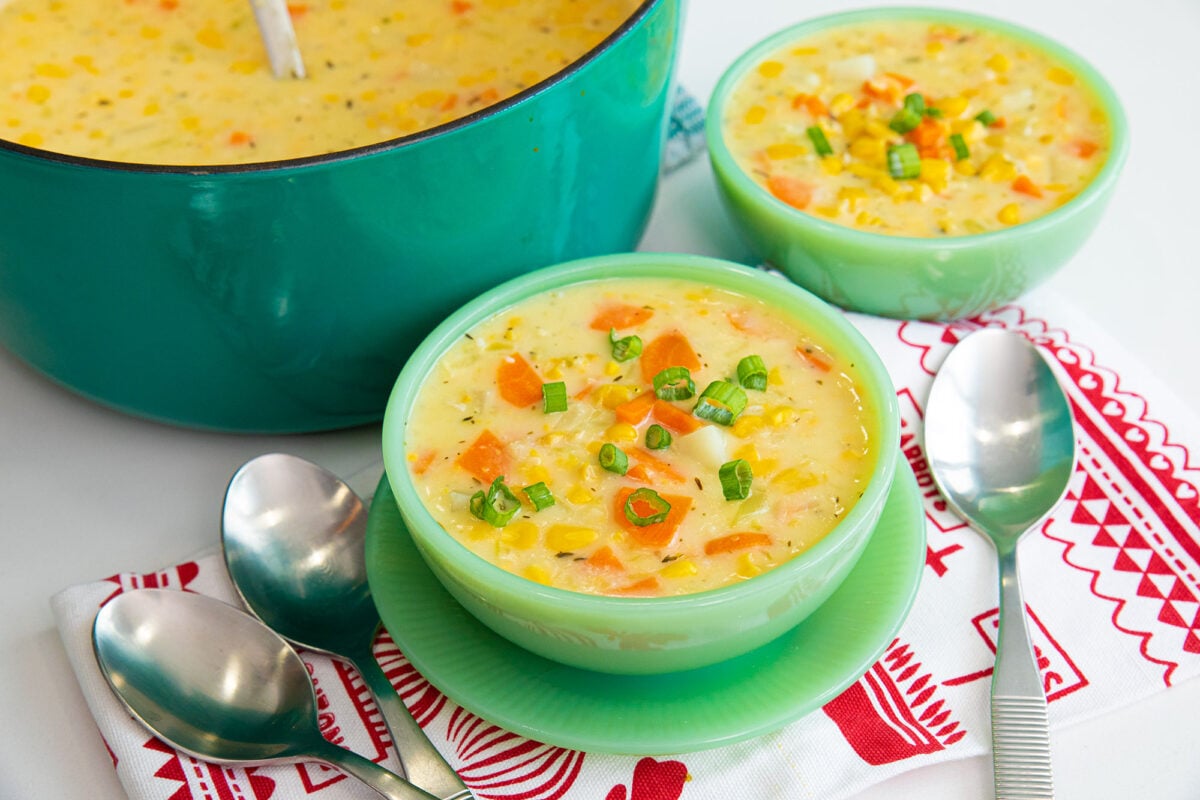Creamy corn soup uses up canned corn and creamed corn, making this a really affordable meal that the entire family will love! Use vegetables from your fridge like carrots, celery and a potato to bulk up this soup and make it a dinner on it's own. #thekitchenmagpie #souprecipes #soup #cornsoup Corn soup in a jadeite bowl with spoons
