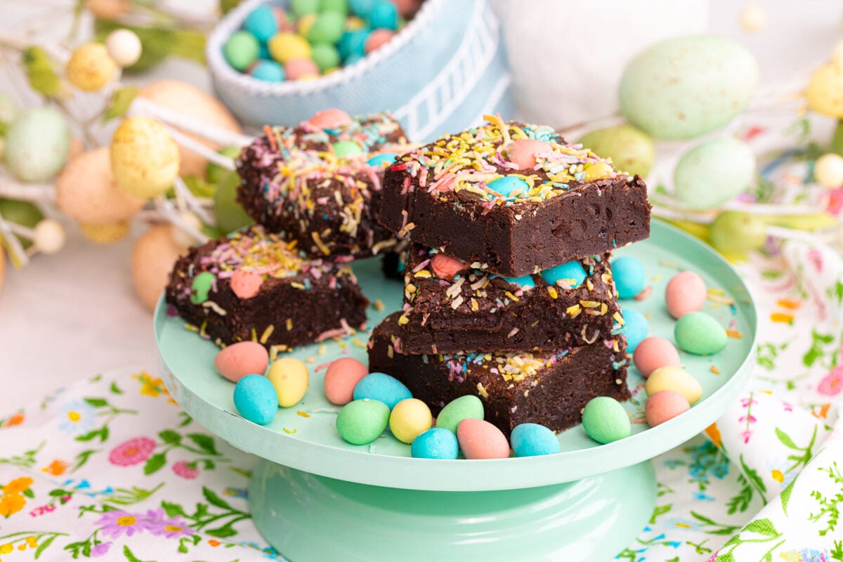 Easter brownies on a stand surrounded by colorful eggs and coconut shavings.