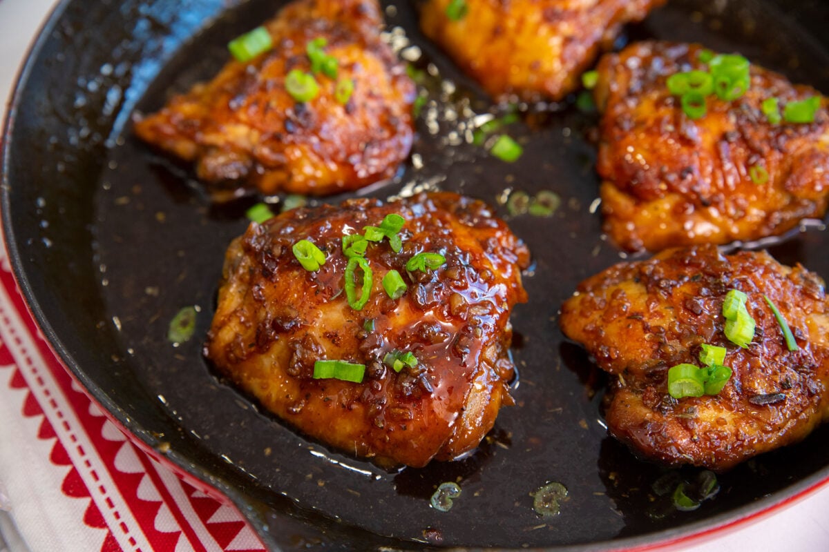 Close-up of garlic brown sugar chicken thighs, pan-seared and glazed, garnished with chopped green onions in a cast-iron skillet filled with savory sauce. The skillet rests on a red and white patterned cloth.