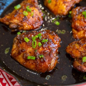 Garlic brown sugar chicken thighs cooked in a skillet with a dark, glossy sauce, garnished with sliced green onions. The dish sits on a red and white patterned cloth.