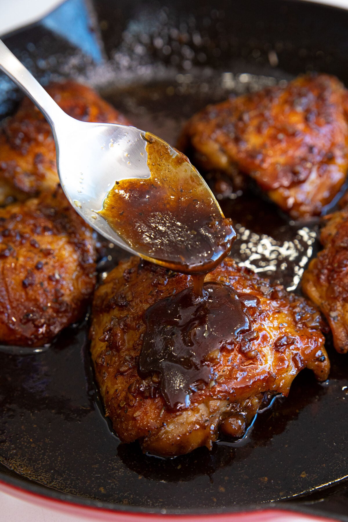A spoon drizzles thick, dark sauce over golden-brown garlic brown sugar chicken thighs sizzling in a skillet.