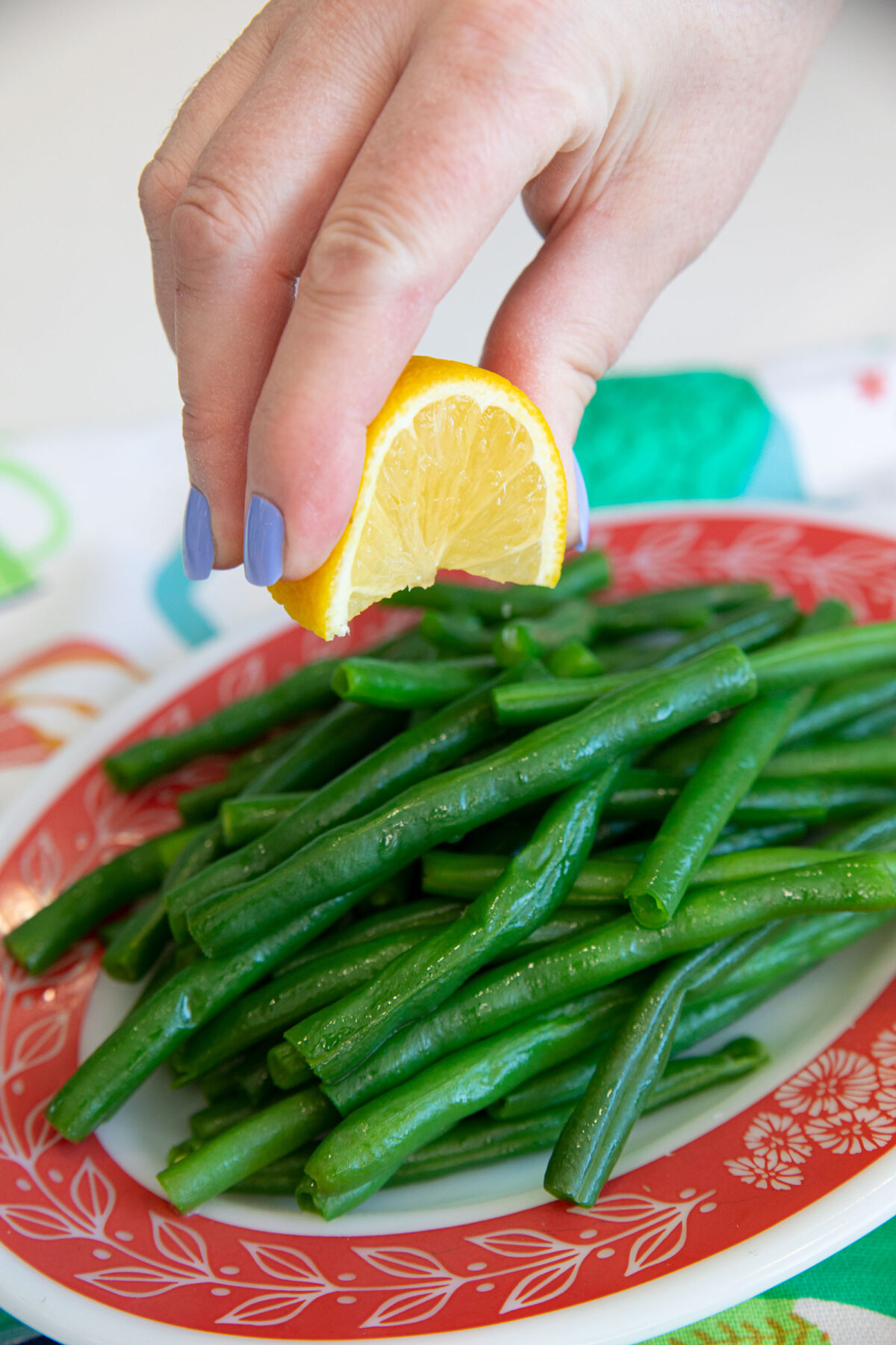 A hand with purple nail polish squeezes a lemon wedge over a plate of cooked green beans on a red and white patterned dish.