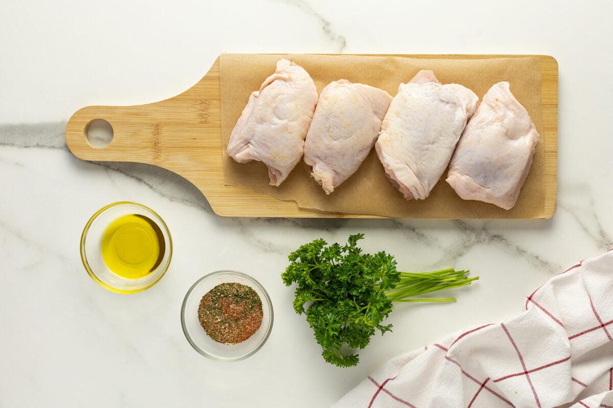 Four raw chicken thighs on a wooden cutting board lined with parchment paper, next to a bowl of olive oil, a bowl of mixed spices, fresh parsley, and a white cloth with red stripes on a marble surface.