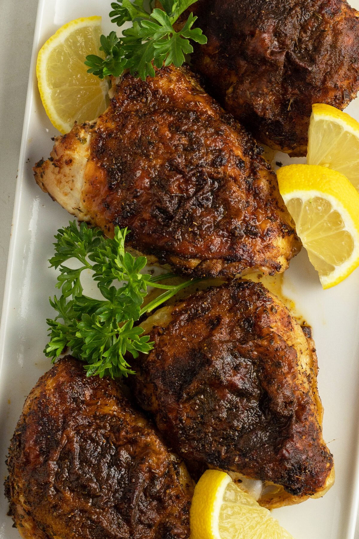 Four pieces of seasoned and roasted chicken garnished with fresh parsley and lemon wedges are arranged on a white rectangular plate.