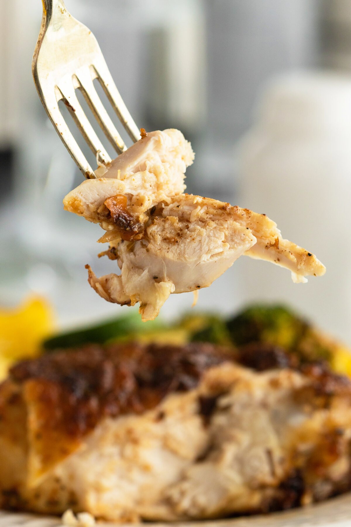 A fork holds a juicy, seasoned piece of cooked chicken above a plate with more chicken and vegetables in the background. The focus is on the fork and chicken, showing its tender texture.