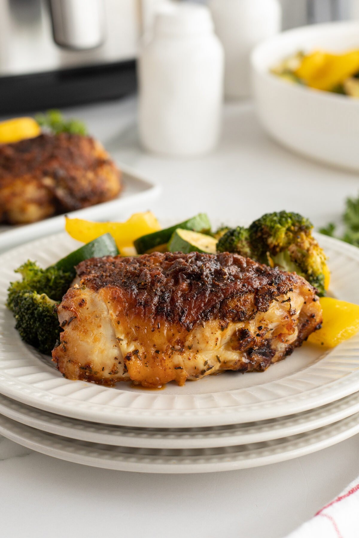 A plate with a piece of seasoned, roasted chicken and a side of sautéed broccoli, zucchini, and yellow bell peppers, served on stacked white plates. Another plate and a bowl of vegetables are in the background.