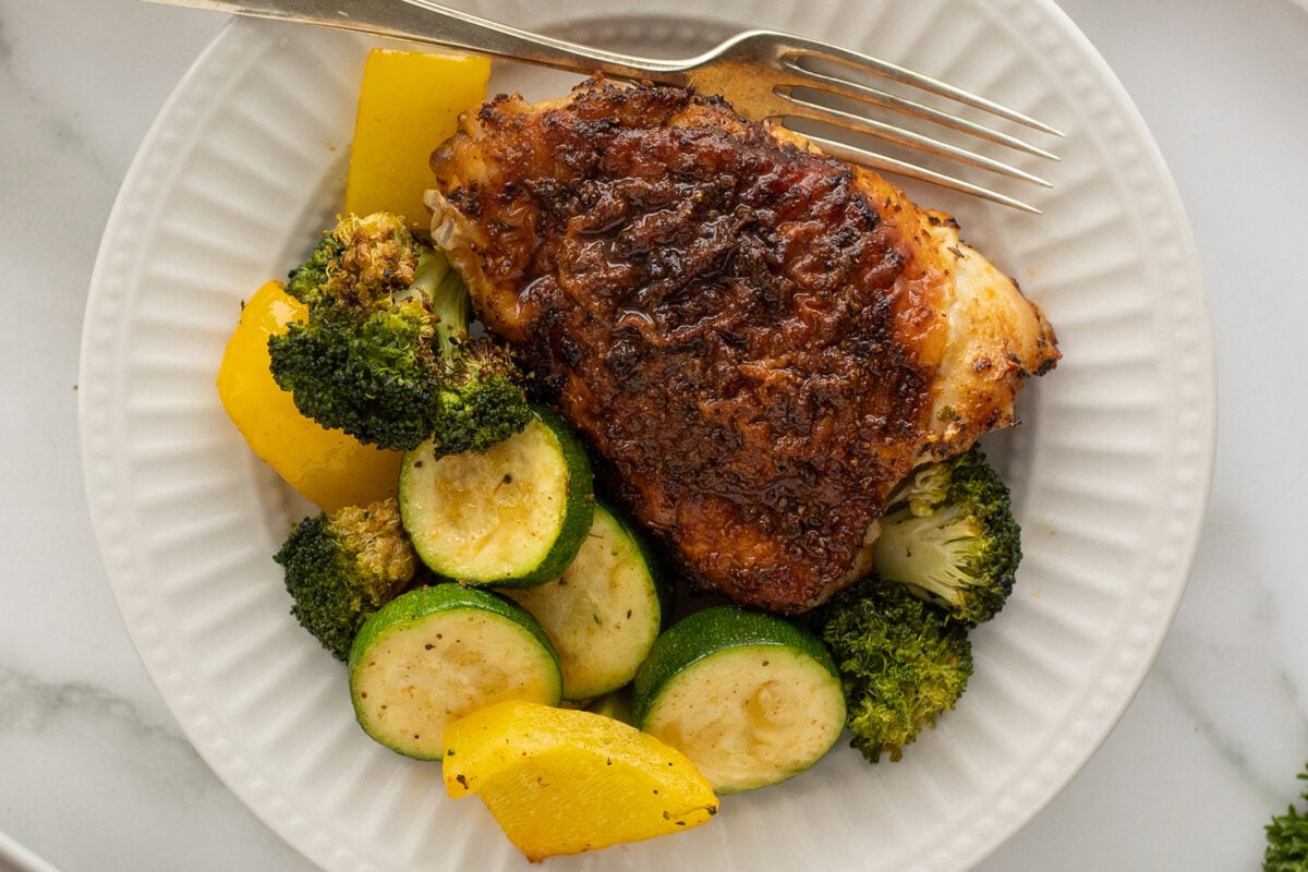 A plate with a seasoned, crispy chicken thigh, accompanied by sautéed zucchini, broccoli, and yellow bell pepper, with a fork resting on the plate.